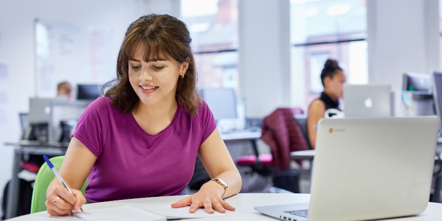 Brunette woman in purple top writing notes on paper with laptop