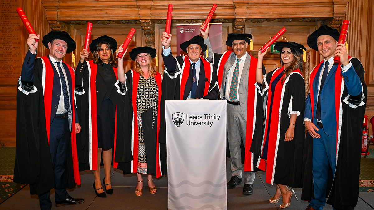 A group of people in graduation caps and gowns, holding up graduation scrolls behind a lectern covered with a white Leeds Trinity University branded cloth.