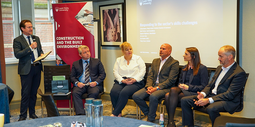 A panel of men and women sitting down in front of a branded Leeds Trinity University banner and having a conversation.
