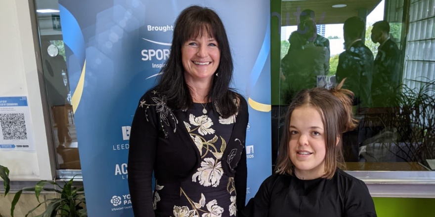 From left to right: brunette woman in floral dress and light brown woman in black dress with glass trophy