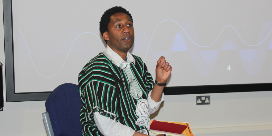Male sits in front of screen wearing black and green striped shawl