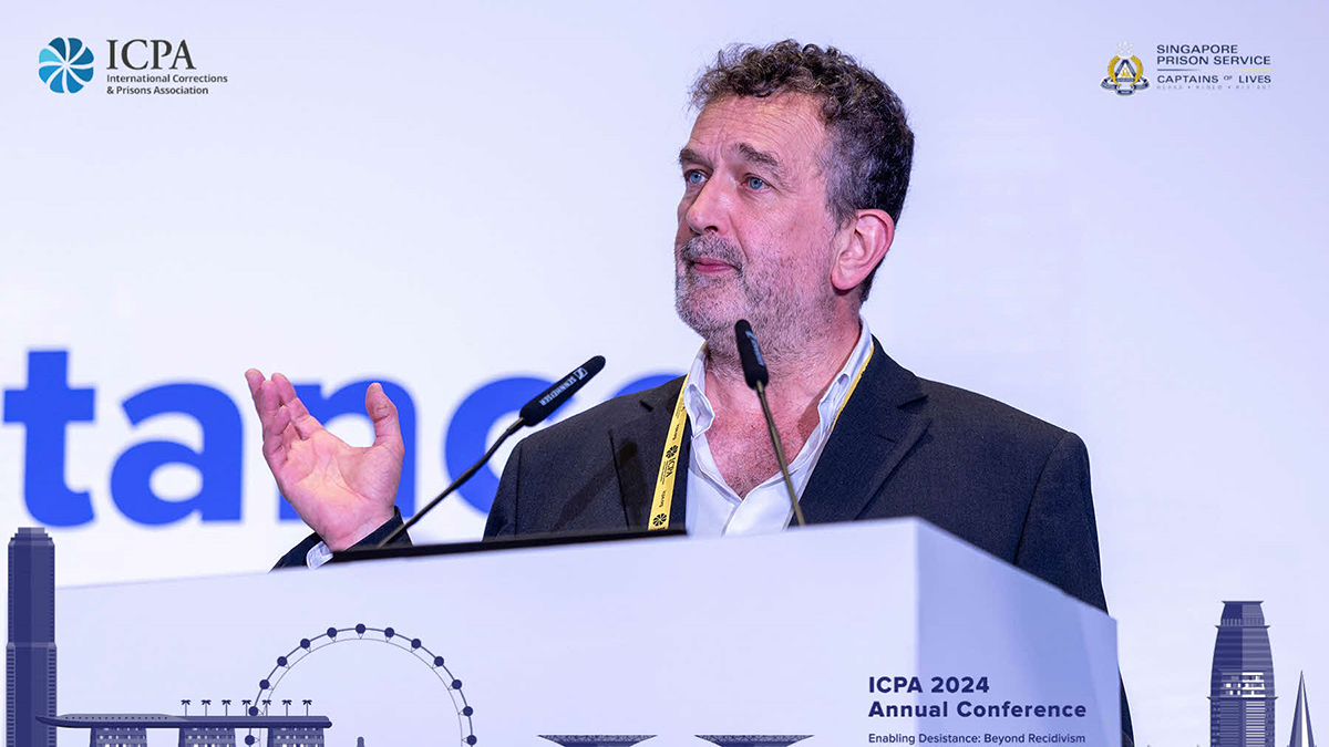 A male academic giving a speech at a conference in Singapore, behind a white lectern and wearing a black blazer.