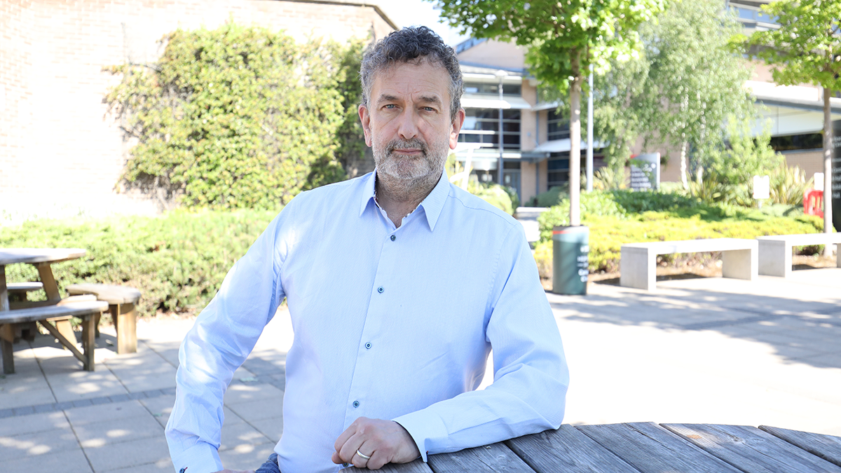 A male academic in a light blue shirt sitting at a wooden table on campus.