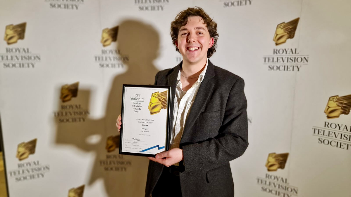 A student in a black suit smiling and holding an award in front of a Royal Television Society backdrop.