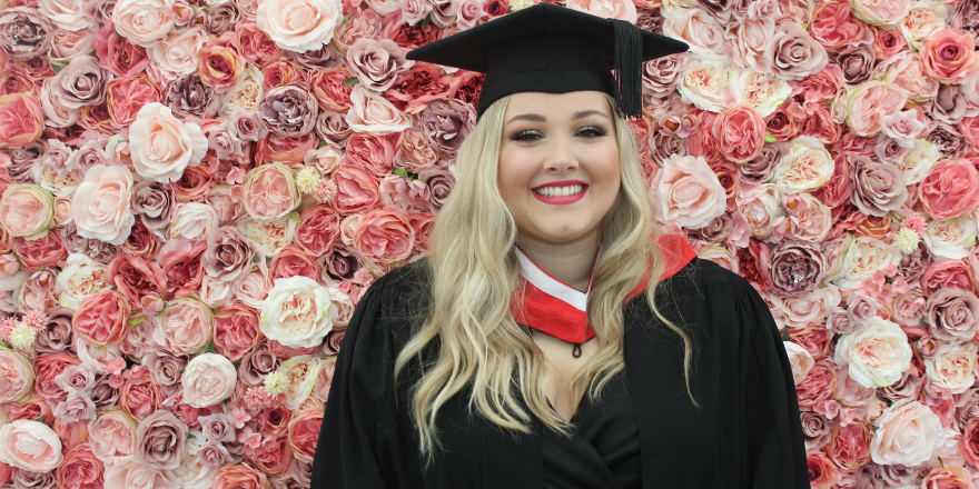Blonde female wearing graduate cap stands in front of flower wall