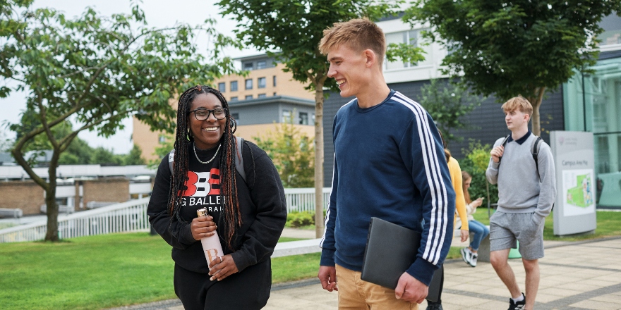 Two students walking and talking at the entrance to campus, with other students and trees in the background