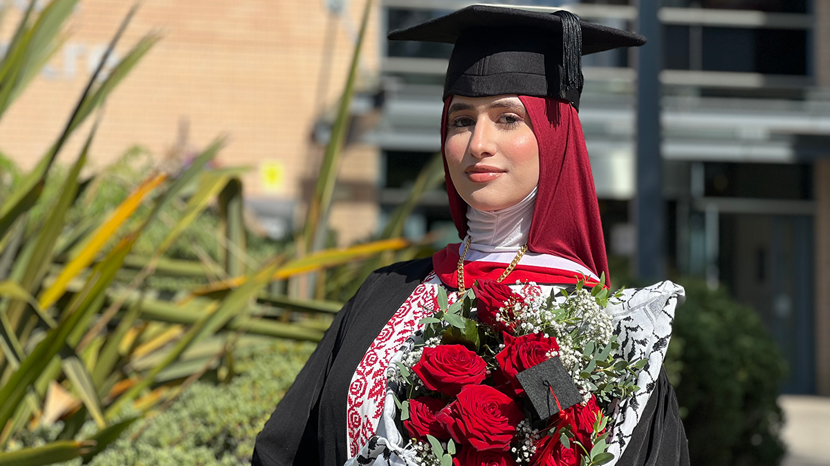 A student in a burgundy hijab, and Graduation cap and gown, holding a bouquet of red roses and smiling.