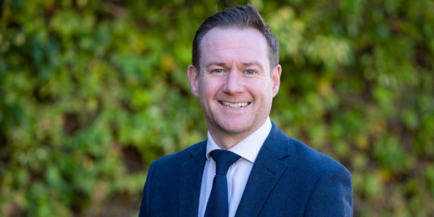 Male with brown hair stands outside wearing dark blue suit and tie