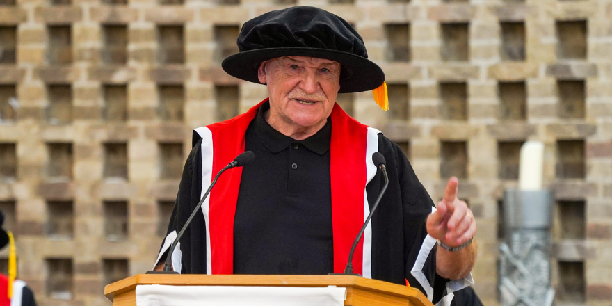 Elderly male wearing cap and gown stands at lectern