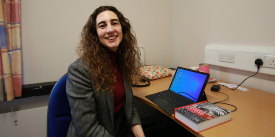 Female with dark curly hair sits at table with laptop and book