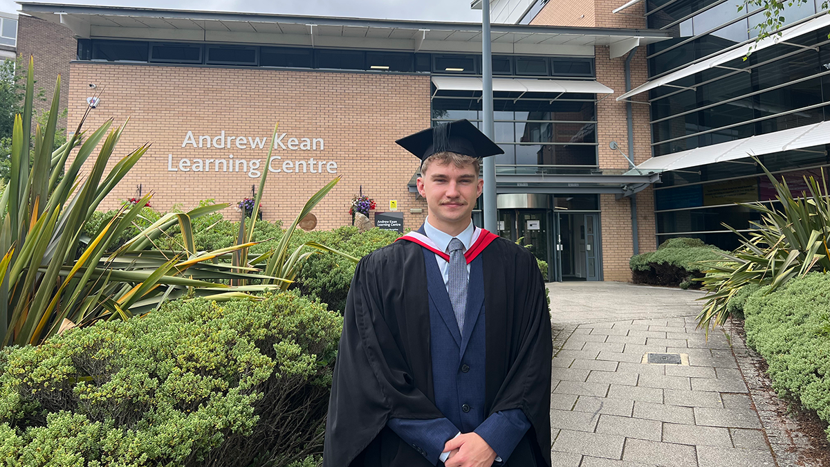 A student in graduation cap and gown smiling in front of the Andrew Kean Learning Centre at Leeds Trinity University.
