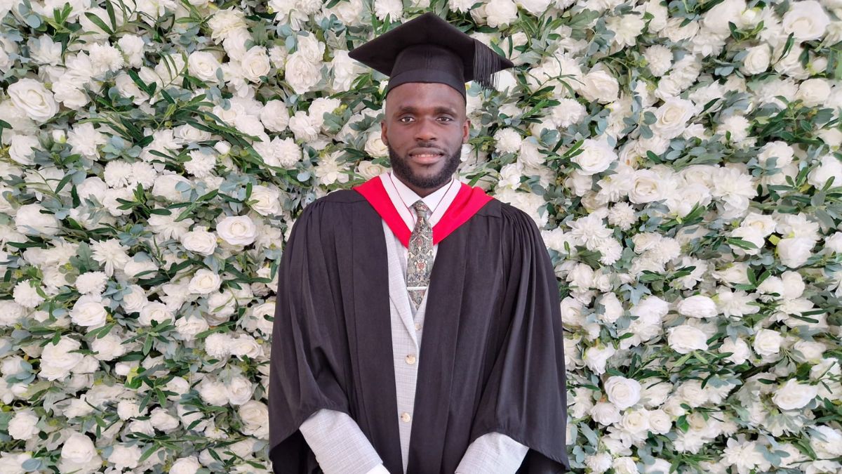 A student wearing a graduation cap and gown stands in front of a flower wall