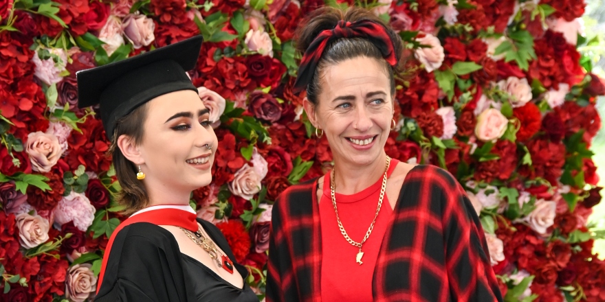 Brunette female graduate in black graduation cap and gown with brunette woman in red top and checked jacket