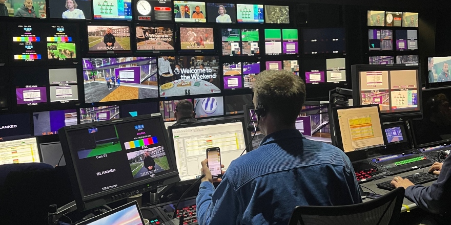A director in a blue shirt sits at a media production desk, watching a number of different screens