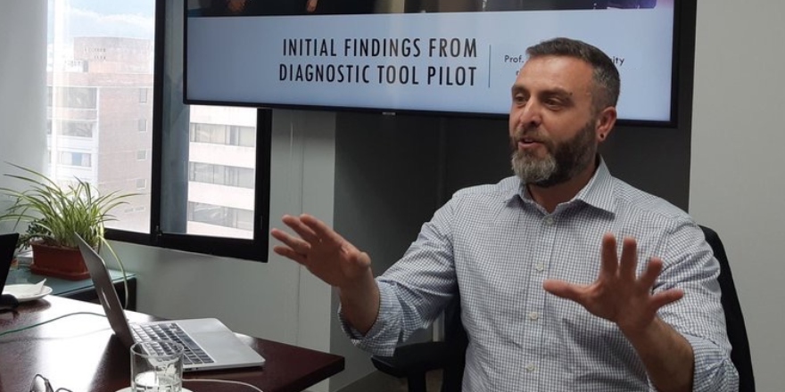 Man with beard sat at a table, actively talking/presenting with a screen behind him
