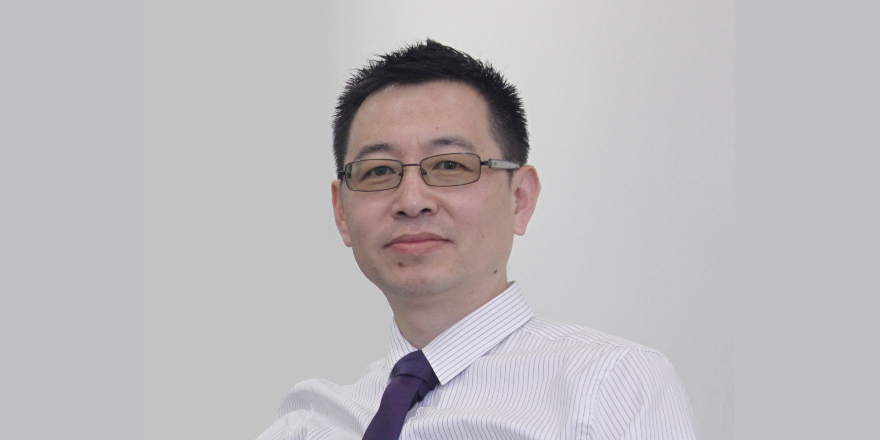 A headshot of a man with brown hair and glasses, sitting in front of a plain white wall