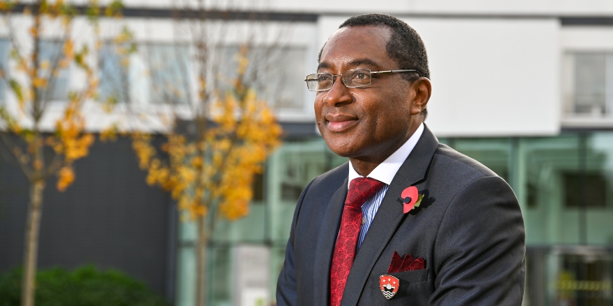 Professor Charles Egbu sat in blue suit with red tie, yellow leaves on tree in background
