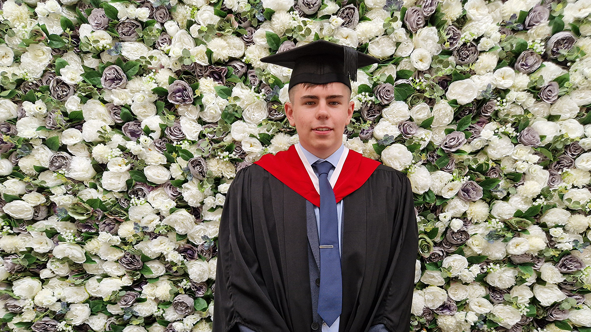 A student in graduation cap and gown smiling in front of a flower wall.
