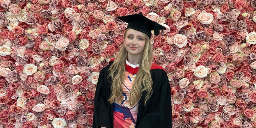 A girl with blonde hair wearing a black graduation cap and gown stands in front of wall of pink flowers