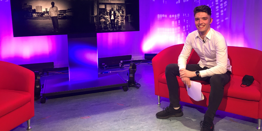 Male with brown hair, white shirt and dark trousers sits on red sofa to the right of tv screen